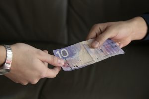 A photograph of one hand exchanging a ten dollar bill with a hand belonging to someone else. 